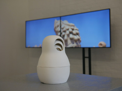 In the foreground is a small white figure modeled after a matryoshka. Behind her, two large television displays show a view of the sky.