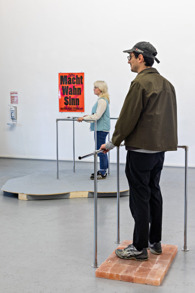 Two people are standing at a distance from each other, each at a railing made of metal bars. Both look straight ahead to the left. The person in front stands on a square of salt stones. The person in the back is standing on a gray wooden plate, which looks like a cloud in its shape.