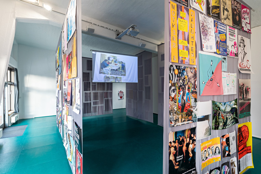 In a high room, gray fabric panels hang from the ceiling and form a room of their own. Many colorful posters are attached to them. In the space in between, a video plays on a small screen.